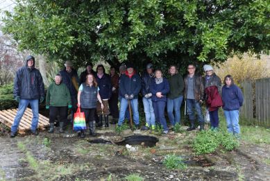 A group of volunteers wearing coats and hats stand together outdoors in a garden area, posing for a photo beside freshly cleared ground and plants.