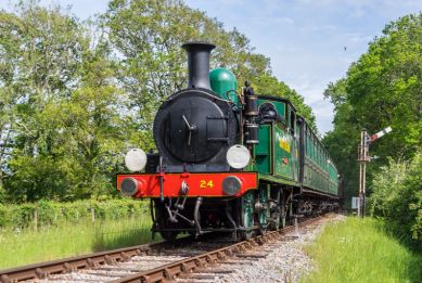 A green vintage steam locomotive travels along a countryside railway line, surrounded by trees and grass on a sunny day.