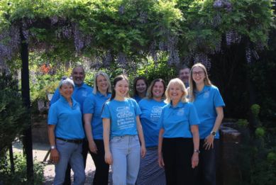 A smiling team of adults wearing matching blue T-shirts stand together on a wooden bridge in a leafy garden setting.
