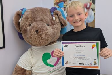 A smiling young boy holds a certificate while standing next to a large teddy bear mascot inside a charity centre.
