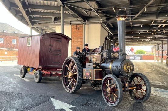 Vintage steam traction engine pulling a wooden wagon under a covered station platform, with two operators seated on the vehicle and small British flags displayed.